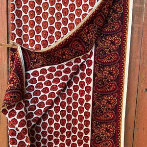 May include: A folded fabric with a repeating pattern of red heart-shaped designs on a white background. A border with paisley and floral patterns in burgundy and gold accents the fabric. The fabric is draped over a wooden rod.