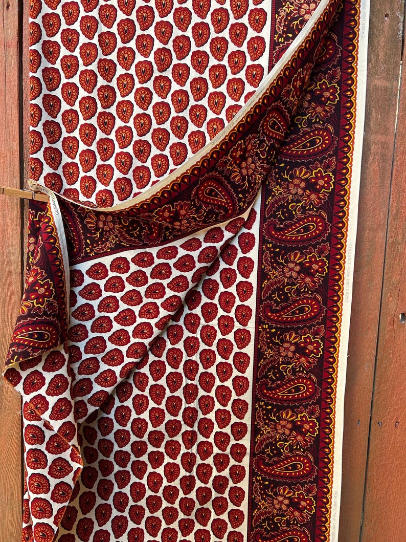 May include: A folded fabric with a repeating pattern of red heart-shaped designs on a white background. A border with paisley and floral patterns in burgundy and gold accents the fabric. The fabric is draped over a wooden rod.