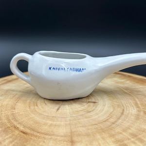 May include: A white ceramic neti pot with a handle and a spout, resting on a wooden surface. The text "KAIVALYADHAM" is printed in blue on the side of the pot. The background is dark.