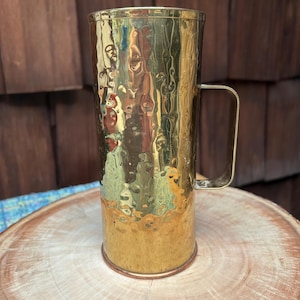 May include: A hammered brass mug with a handle. The mug has a cylindrical shape and a shiny, textured surface. The mug is sitting on a wooden surface.