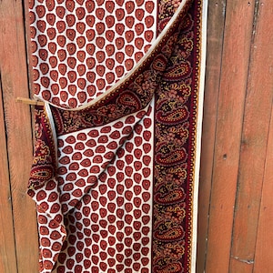 May include: A folded fabric with a white background and a repeating pattern of red teardrop shapes. A burgundy paisley border and trim are visible. The fabric is hanging on a wooden fence.
