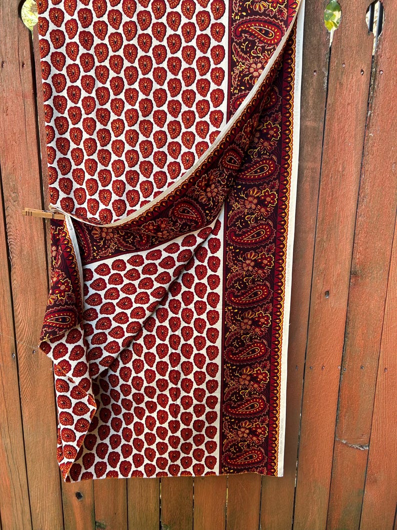 May include: A folded fabric with a white background and a repeating pattern of red teardrop shapes. A burgundy paisley border and trim are visible. The fabric is hanging on a wooden fence.