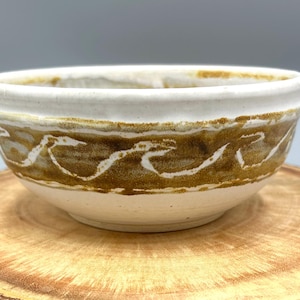 May include: A handmade ceramic bowl with a white interior and a decorative band of brown and white wave-like patterns around the exterior. The bowl is set on a wooden surface.