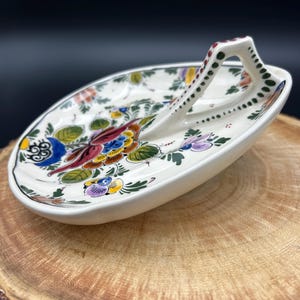 May include: Oval-shaped ceramic serving dish with a handle, featuring a hand-painted floral design in vibrant colors. The dish has a white base with a black rim and is set on a wooden surface. The handle is decorated with green and red dots.