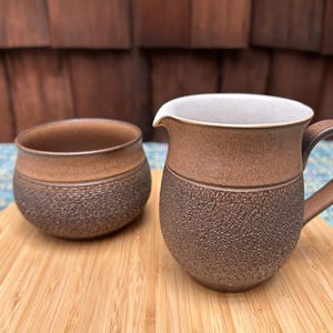 May include: A brown and white ceramic sugar bowl and creamer set with a textured finish. The set is made of stoneware and has a rustic look.