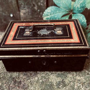 May include: A vintage black metal cash box with a handle and a lock. The box features a gold and red border design. The words "ENGLISH MAKE" are visible on the front. The box is likely used for storing money or valuables.