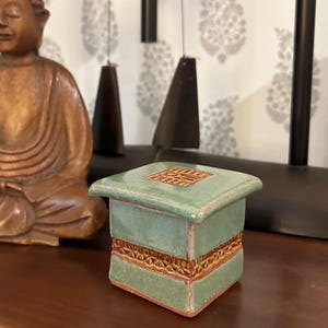 May include: A small, square, ceramic box with a light green glaze. The box features a decorative band of brown, textured detailing around the middle and on the lid. The box is sitting on a dark brown surface.