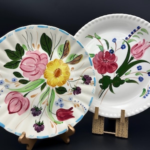 May include: Two white ceramic plates with hand-painted floral designs. One plate has a ruffled edge with pink, yellow, and purple flowers. The other plate has a beaded edge with red and pink flowers. Both plates are on display stands.
