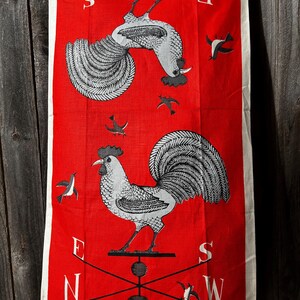 May include: A red kitchen towel with a rooster and weather vane design. The towel features two roosters in black and white, with a weather vane and compass points. Small bird illustrations are scattered around the roosters.