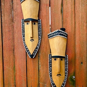 May include: Two wooden tribal masks with elongated faces and geometric patterns. The masks are painted in natural wood tones with black and white accents. They are mounted on a weathered, reddish-brown wooden fence.