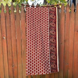 May include: A rectangular textile with a white background and a repeating pattern of red heart-shaped designs. The right side features a burgundy border with a paisley pattern. The textile is hanging on a wooden rod against a wooden fence.
