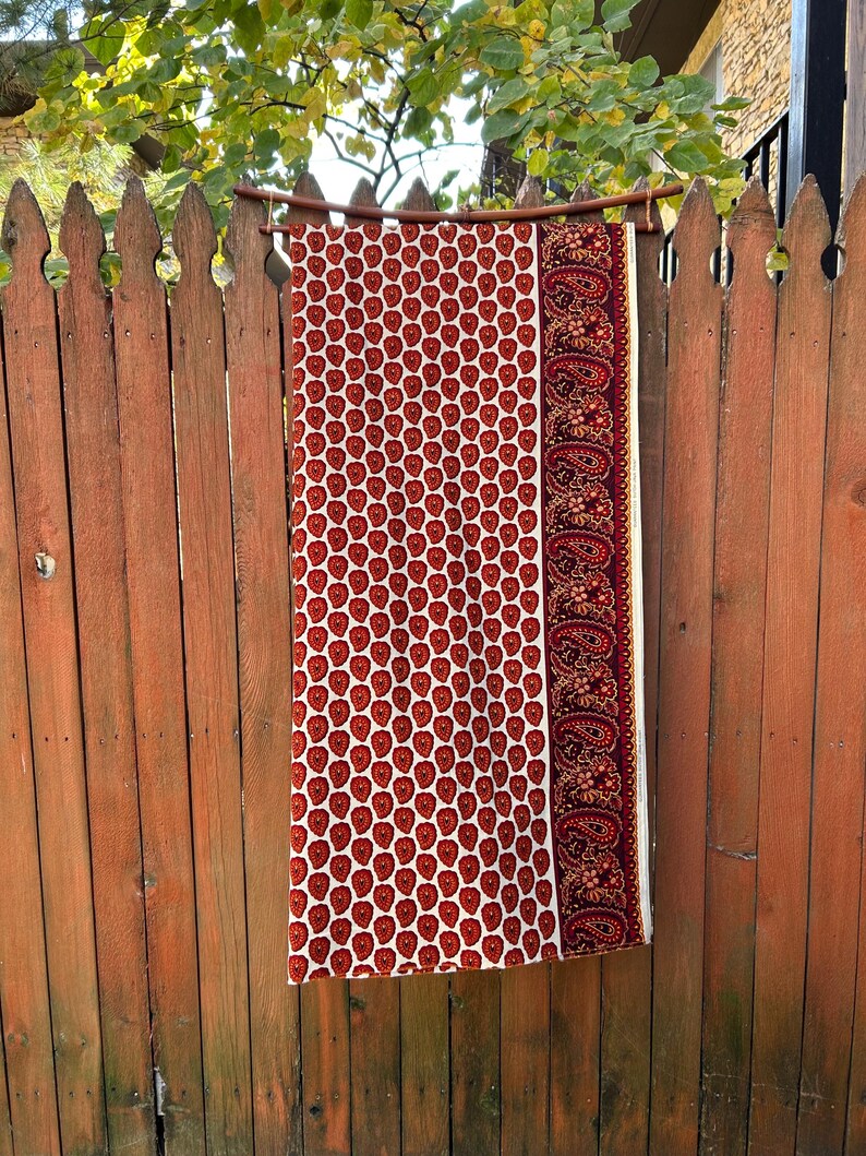 May include: A rectangular textile with a white background and a repeating pattern of red heart-shaped designs. The right side features a burgundy border with a paisley pattern. The textile is hanging on a wooden rod against a wooden fence.