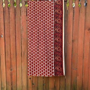 May include: A rectangular fabric with a repeating red apple pattern on a cream background. A burgundy paisley border is on the right side. The fabric is hanging on a wooden rod against a wooden fence.