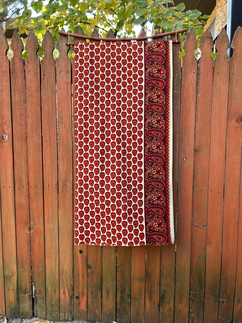 May include: A rectangular fabric with a repeating red apple pattern on a cream background. A burgundy paisley border is on the right side. The fabric is hanging on a wooden rod against a wooden fence.