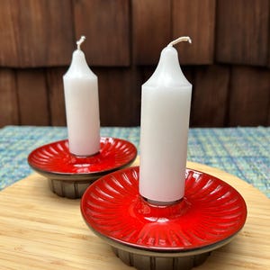 May include: Two red ceramic candle holders with white candles. The holders have a fluted design and a dark brown base.