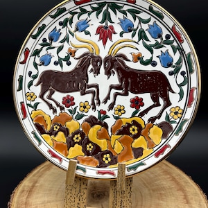 May include: A decorative ceramic plate featuring two brown goats facing each other. The plate has a white background with floral and leaf designs in blue, red, yellow, and green. The rim is gold.