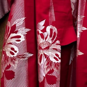 May include: Close-up of a red kimono with silver floral embroidery. The fabric has a subtle, textured pattern in the background. The design features large, stylized flowers and leaves, creating a striking contrast against the vibrant red.