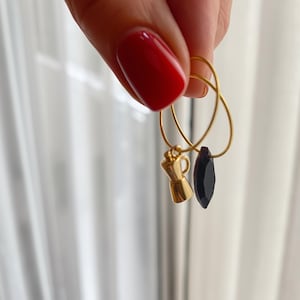 May include: Gold hoop earrings with charms. One charm is a gold coffee maker, and the other is a black, teardrop-shaped gem. The earrings are held up by a hand with red nail polish.