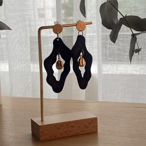 Op de afbeelding: Een paar donkerblauwe, abstract gevormde oorbellen met gouden accenten. De oorbellen worden gepresenteerd op een goudkleurige standaard met een houten voet. De oorbellen hebben een getextureerd oppervlak en een kleine, bungelende gouden kraal.