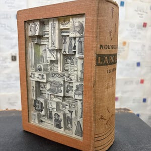 以下が含まれることがあります： ベージュの布製カバーに「NOUVEAU LAROUSSE ILLUSTRE」と記された、中をくり抜いたヴィンテージ本。内部の空洞には、それぞれ異なる白黒の線画が描かれた小さな長方形の木製ブロックが密集して詰め込まれています。
