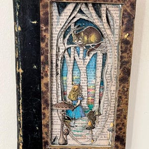 May include: An altered book featuring a layered paper art scene from Alice in Wonderland. The artwork includes Alice, the Cheshire Cat, and whimsical mushrooms, set against a backdrop of book pages and a blue sky.