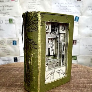 May include: A handcrafted, green-covered book with a detailed diorama inside, featuring miniature sewing tools and illustrations. The book's spine has gold accents and the word "NEEDLEWORK". The background shows vintage envelopes.