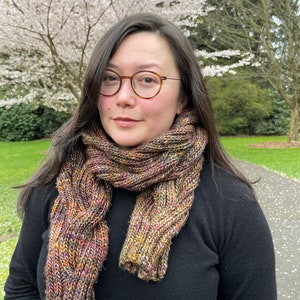 May include: A chunky, hand-knit scarf featuring a variegated yarn blend of earthy tones including brown, gold, green, and hints of purple. The scarf displays a prominent vertical cable knit pattern and is worn wrapped around the neck over a black sweater.