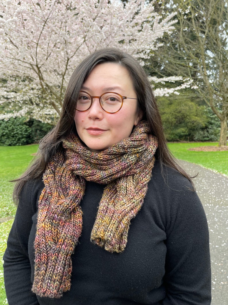 May include: A chunky, hand-knit scarf featuring a variegated yarn blend of earthy tones including brown, gold, green, and hints of purple. The scarf displays a prominent vertical cable knit pattern and is worn wrapped around the neck over a black sweater.