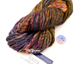 May include: A skein of super bulky Rasta yarn in the 862 Piedras colorway, featuring a variegated blend of purple, orange, olive green, and brown tones. The yarn label specifies 100% merino wool, 90 yards, and a gauge of 8-10 stitches per 4 inches.