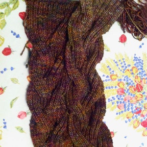 May include: A hand-knitted scarf in progress featuring a complex cable stitch pattern. The yarn is a variegated blend of deep brown, dark purple, forest green, and burnt orange tones. A single knitting needle remains inserted at the top edge.