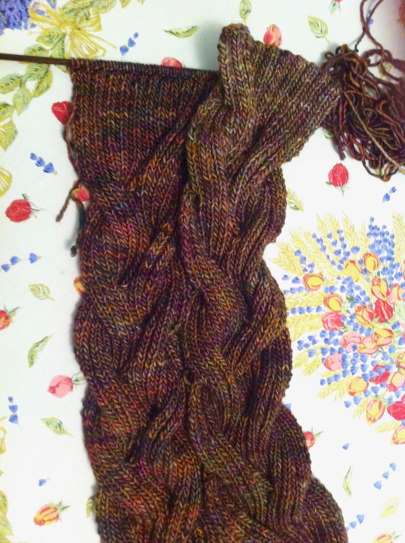 May include: A hand-knitted scarf in progress featuring a complex cable stitch pattern. The yarn is a variegated blend of deep brown, dark purple, forest green, and burnt orange tones. A single knitting needle remains inserted at the top edge.