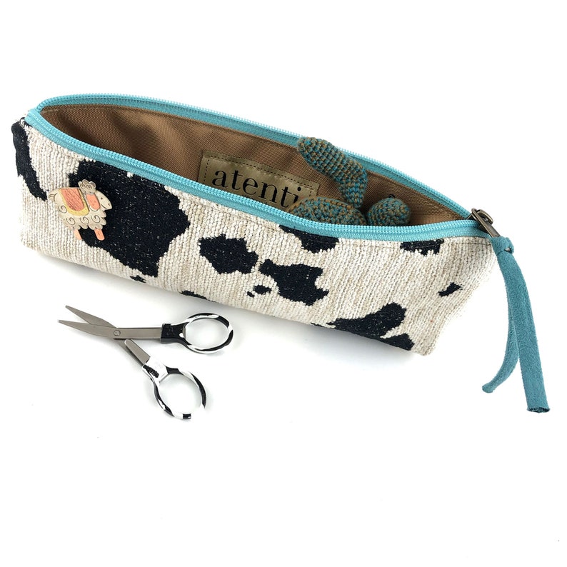 Pode incluir: Uma bolsa de z&iacute;per com estampa de vaca branca e preta com um puxador de z&iacute;per turquesa. A bolsa est&aacute; aberta e cont&eacute;m um pequeno brinquedo de croch&ecirc;. O texto "atenTi" &eacute; vis&iacute;vel no interior da bolsa.