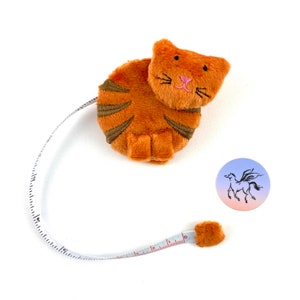 May include: A plush orange tabby cat-shaped tape measure with a white tape and black markings. The tape measure is curled into a circle with the cat's head at the top. The cat has pink embroidered nose and whiskers.