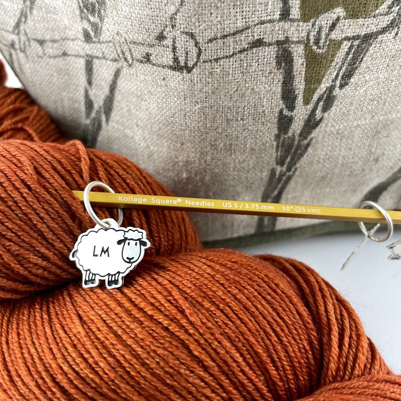 May include: A skein of rust-colored yarn with a sheep-shaped stitch marker attached to a knitting needle. The needle is marked "Kollage Square Needles US 5 / 3.75 mm 10" (25 cm)." The stitch marker has the letters "LM" on it.
