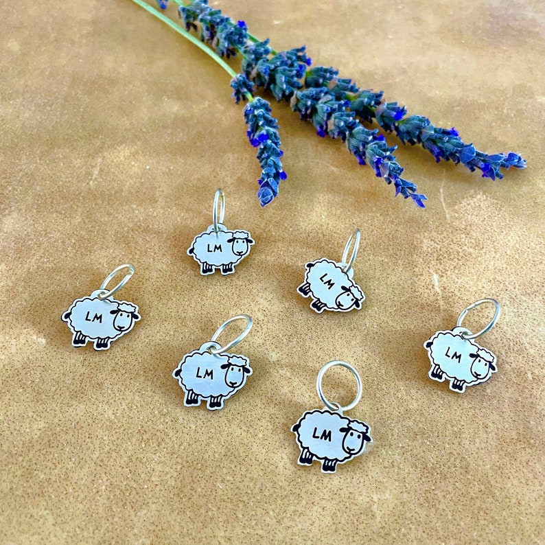 Six silver colored metal sheep stitch markers each hang on a small ring. Stitch markers have a black outline accentuating the fleece and drawing out a face, feet and LM in the center of the body.