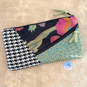May include: A zippered pouch featuring a black fabric panel with a colorful floral pattern, a black and white houndstooth section, and a sage green panel with blue stitched details. The pouch includes two light green suede zipper pulls.