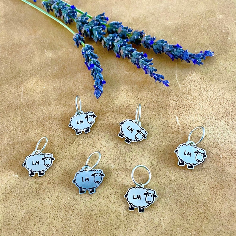 May include: Six silver-toned sheep-shaped stitch markers with the letters "LM" on their bodies. The sheep are white with black outlines and legs. A sprig of lavender with purple flowers is in the background. The markers are on a brown surface.