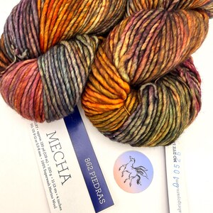 May include: A skein of Mecha yarn in the color 862 Piedras, featuring a variegated blend of orange, purple, and green tones. The label indicates 100g, 130 yards, and 100% Superwash Merino Wool, with a circular sticker showing a horse illustration.