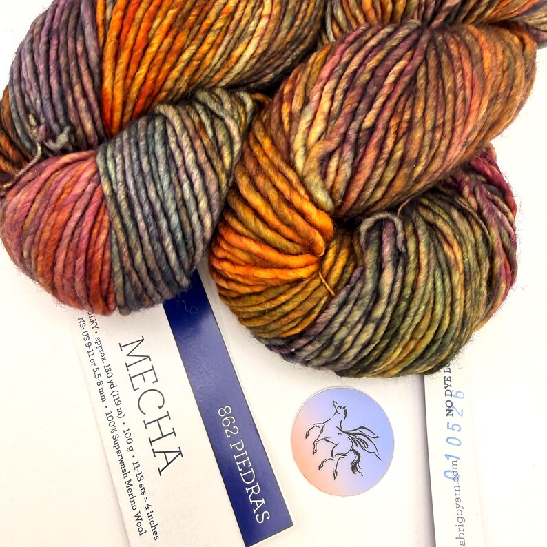 May include: A skein of Mecha yarn in the color 862 Piedras, featuring a variegated blend of orange, purple, and green tones. The label indicates 100g, 130 yards, and 100% Superwash Merino Wool, with a circular sticker showing a horse illustration.