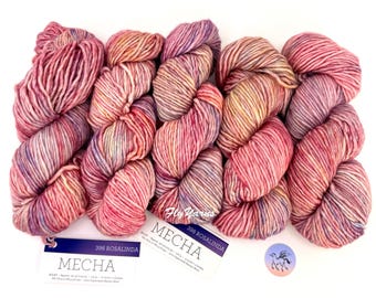 Puede incluir: Cinco madejas de hilo matizado en tonos rosa, naranja y morado. Cada etiqueta dice MECHA 398 ROSALINDA, indicando 100g y 130 yardas. Una pequeña pegatina circular con el logotipo de un caballo alado se encuentra junto al hilo.