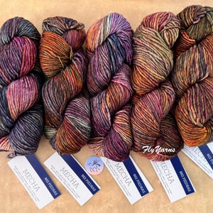 May include: Five skeins of Mecha 862 Piedras yarn, each featuring a variegated blend of deep purple, orange, brown, and charcoal tones. Each skein is accompanied by a white rectangular label with blue text detailing the product specifications.