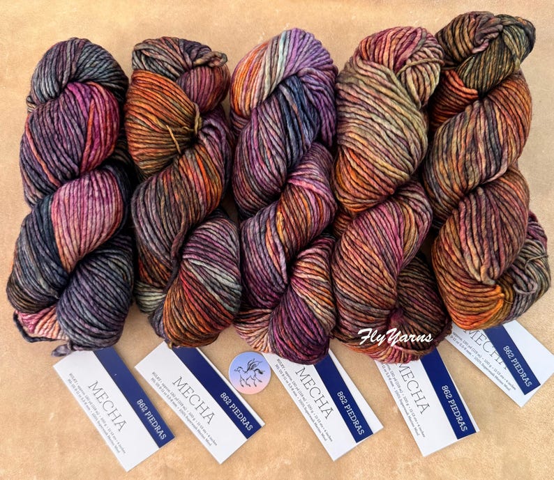 May include: Five skeins of Mecha 862 Piedras yarn, each featuring a variegated blend of deep purple, orange, brown, and charcoal tones. Each skein is accompanied by a white rectangular label with blue text detailing the product specifications.