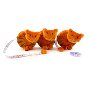 May include: Three orange plush cat-shaped measuring tapes with a white background. The cats are curled up with their tails forming the tape measure.