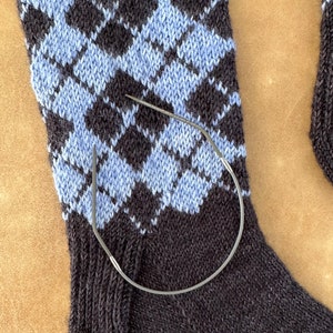 May include: A close-up of a blue and black knitted sock with a diamond pattern. A metal knitting needle is wrapped around the sock.