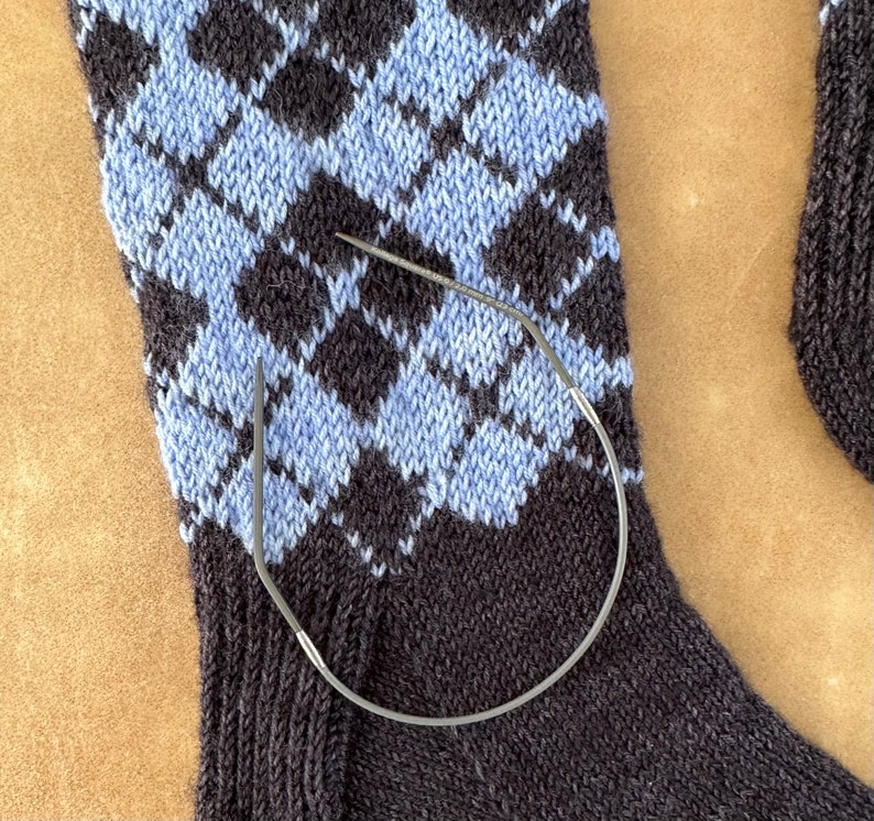 May include: A close-up of a blue and black knitted sock with a diamond pattern. A metal knitting needle is wrapped around the sock.