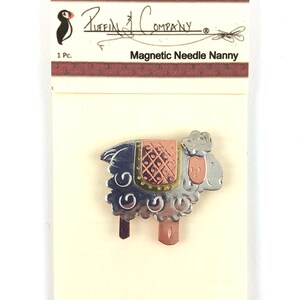 Pode incluir: Um porta-agulhas magn&eacute;tico de prata e cobre em forma de ovelha. A ovelha tem uma sela com padr&atilde;o rosa e dourado. A embalagem diz "Puffin Company" e "Magnetic Needle Nanny".