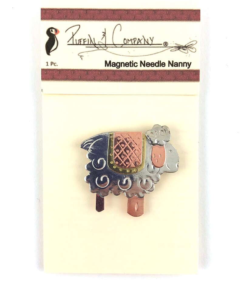 Pode incluir: Um porta-agulhas magn&eacute;tico de prata e cobre em forma de ovelha. A ovelha tem uma sela com padr&atilde;o rosa e dourado. A embalagem diz "Puffin Company" e "Magnetic Needle Nanny".