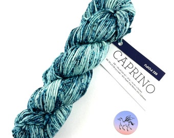 Puede incluir: Un ovillo de hilo Caprino en tonos azules y verde azulado. El hilo está etiquetado con el nombre "CAPRINO" y detalles que incluyen el peso, la longitud y el contenido de fibra. También es visible una pequeña pegatina circular con una ilustración de un pegaso.