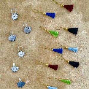 May include: An assortment of knitting stitch markers and tassels. The stitch markers feature a sheep design with the initials "LM". The tassels come in various colors, including blue, green, and burgundy, with gold-colored metal loops.