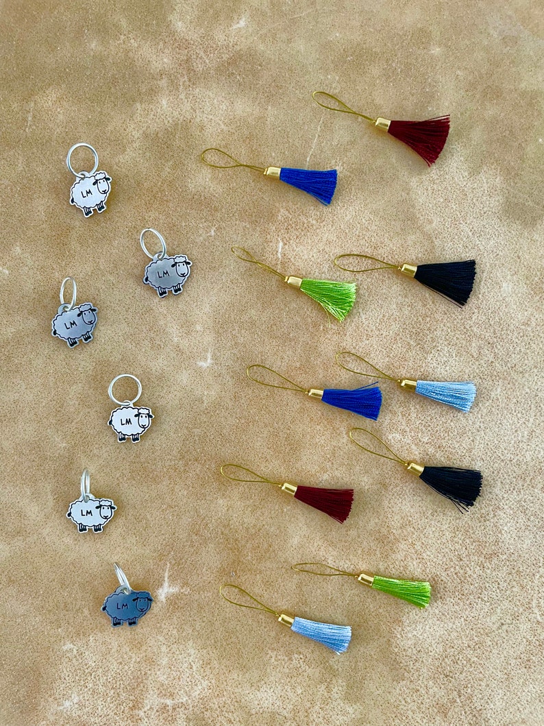 May include: An assortment of knitting stitch markers and tassels. The stitch markers feature a sheep design with the initials "LM". The tassels come in various colors, including blue, green, and burgundy, with gold-colored metal loops.